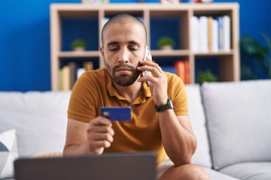 Young latin man talking on smartphone holding credit card with serious expression at home