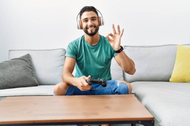 Handsome hispanic man wearing headphones playing video game holding controller doing ok sign with fingers, smiling friendly gesturing excellent symbol 
