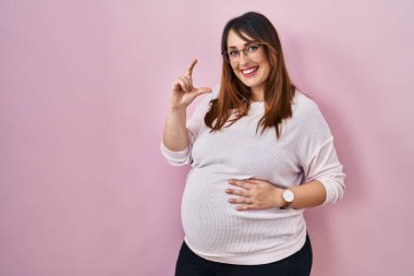 Pregnant woman standing over pink background smiling and confident gesturing with hand doing small size sign with fingers looking and the camera. measure concept. 