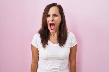 Middle age brunette woman standing over pink background angry and mad screaming frustrated and furious, shouting with anger. rage and aggressive concept. 