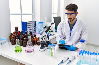 Young hispanic man wearing scientist uniform writing on clipboard working at laboratory