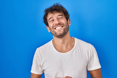 Hispanic young man standing over blue background hands together and fingers crossed smiling relaxed and cheerful. success and optimistic 