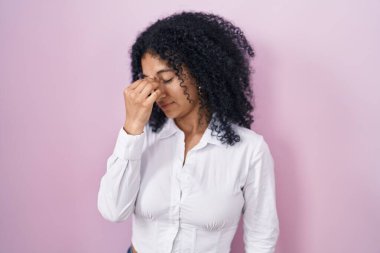 Hispanic woman with curly hair standing over pink background tired rubbing nose and eyes feeling fatigue and headache. stress and frustration concept. 