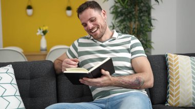 Young caucasian man reading a book sitting on the sofa at home