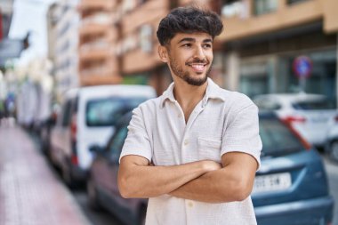 Genç Arap adam gülümsüyor. Kollarını kavuşturup caddede duruyor.
