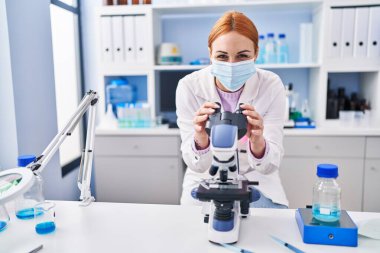 Laboratuvarda mikroskop kullanarak tıbbi maske takan beyaz kadın bilim adamı.