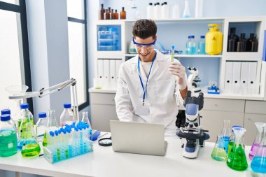 Laboratuvarda dizüstü bilgisayarda test tüpü tutan bilim adamı üniforması giyen İspanyol genç adam.