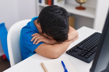 Adorable hispanic boy student using computer with stressed expression at classroom