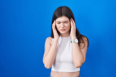 Young caucasian woman standing over blue background with hand on head, headache because stress. suffering migraine. 