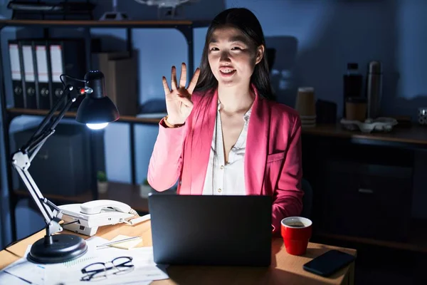 Chinese young woman working at the office at night showing and pointing up with fingers number four while smiling confident and happy.