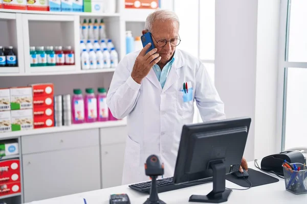 薬局でパソコンを使ってスマホで話すシニア白髪の男性薬剤師 — ストック写真