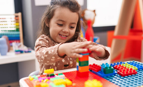 Adorable hispanic girl playing with construction blocks sitting on table at kindergarten