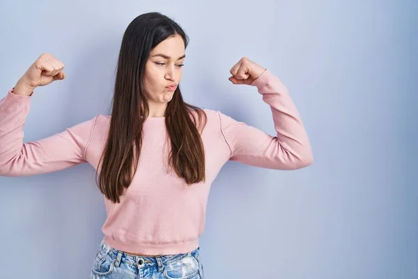 Young brunette woman standing over blue background showing arms muscles smiling proud. fitness concept.