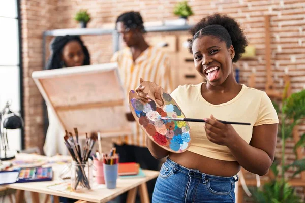 Young black painter woman at art studio holding palette sticking tongue out happy with funny expression.