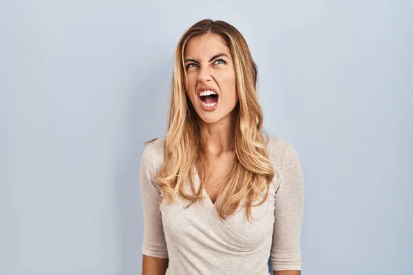 Young blonde woman standing over isolated background angry and mad screaming frustrated and furious, shouting with anger. rage and aggressive concept.