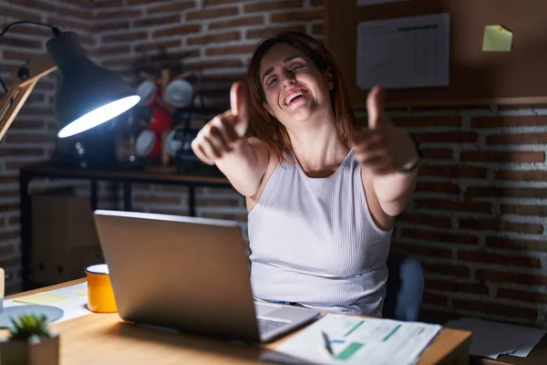 Brunette woman working at the office at night approving doing positive gesture with hand, thumbs up smiling and happy for success. winner gesture.