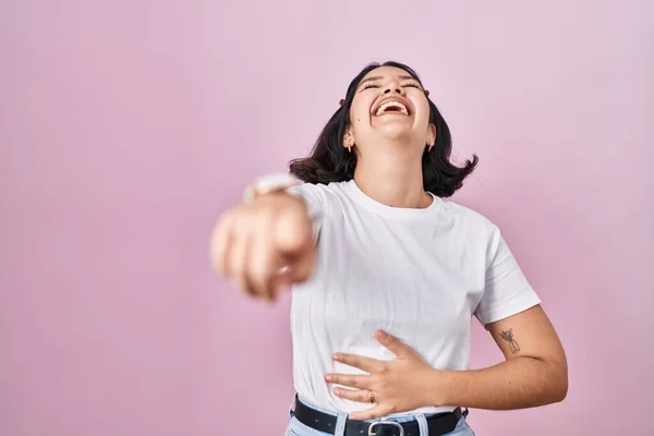 Young hispanic woman wearing casual white t shirt over pink background laughing at you, pointing finger to the camera with hand over body, shame expression