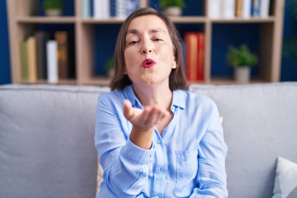 Middle age hispanic woman sitting on the sofa at home looking at the camera blowing a kiss with hand on air being lovely and sexy. love expression.