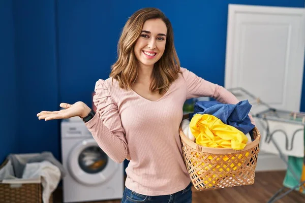 Young woman holding laundry basket smiling cheerful presenting and pointing with palm of hand looking at the camera.