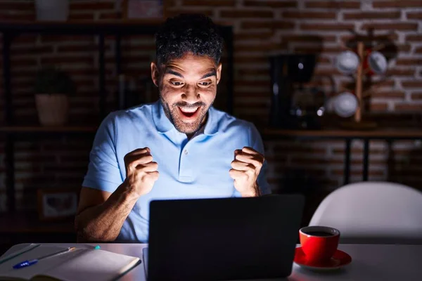 Hispanic man with beard using laptop at night celebrating surprised and amazed for success with arms raised and open eyes. winner concept.