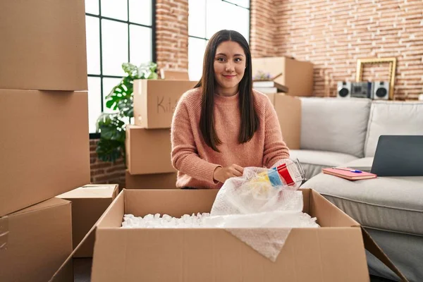 Young hispanic woman unpacking cardboard box sitting on floor at new home