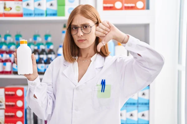 Young redhead woman working at pharmacy drugstore holding sun screen with angry face, negative sign showing dislike with thumbs down, rejection concept