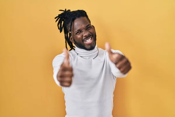 黄色の背景の上にタートルネックセーターを身に着けている恐ろしいロックを持つアフリカの男は 手で積極的なジェスチャーを行うことを承認し 親指を笑顔と成功のために幸せ 勝者のジェスチャー — ストック写真