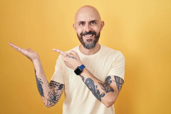 Hispanic man with tattoos standing over yellow background amazed and smiling to the camera while presenting with hand and pointing with finger.