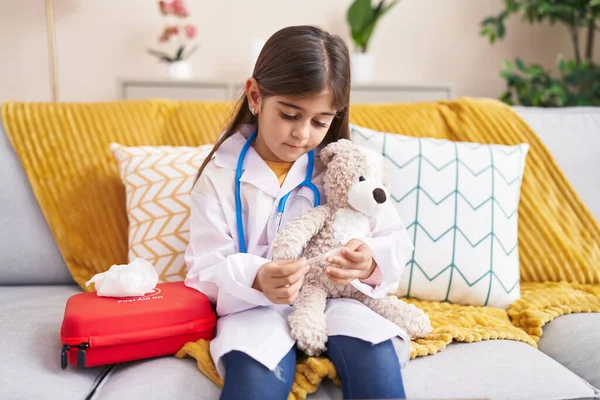 可爱的惊慌失措的女孩穿着医生制服在家里包扎泰迪熊的胳膊 — 图库照片