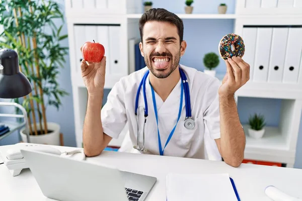 Young Hispanic Dietitian Man Holding Doughnut Apple Sticking Tongue Out — Stockfoto