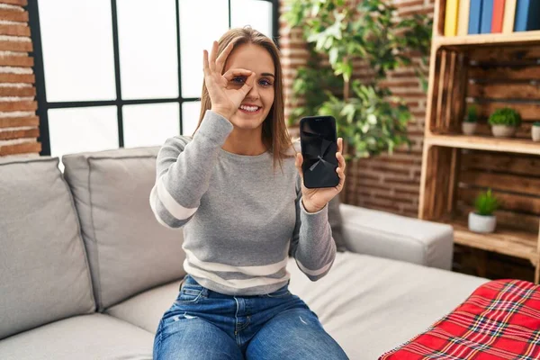Young woman holding broken smartphone showing cracked screen smiling happy doing ok sign with hand on eye looking through fingers