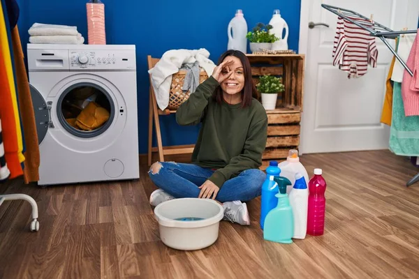 Young hispanic woman doing laundry washing by hand smiling happy doing ok sign with hand on eye looking through fingers