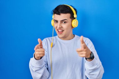 Non binary person listening to music using headphones pointing fingers to camera with happy and funny face. good energy and vibes. 