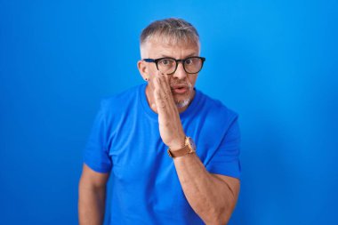 Hispanic man with grey hair standing over blue background hand on mouth telling secret rumor, whispering malicious talk conversation 