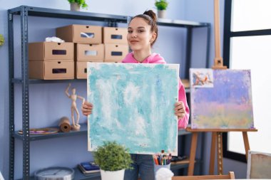 Young hispanic girl holding canvas winking looking at the camera with sexy expression, cheerful and happy face. 