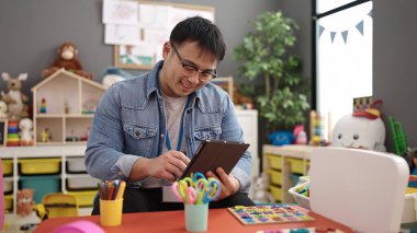 Young chinese man preschool teacher using touchpad sitting on table at kindergarten