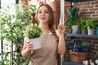 Young caucasian woman working at florist shop holding plant smiling with an idea or question pointing finger with happy face, number one 