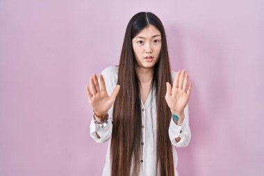 Chinese young woman standing over pink background moving away hands palms showing refusal and denial with afraid and disgusting expression. stop and forbidden. 