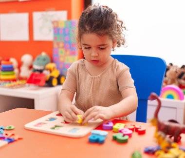 Adorable hispanic girl playing with maths puzzle game sitting on table at kindergarten