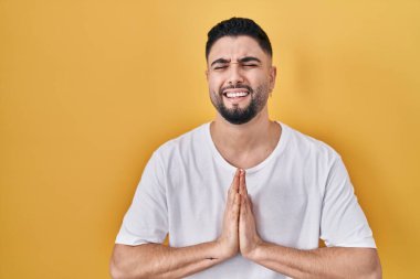 Young handsome man wearing casual t shirt over yellow background begging and praying with hands together with hope expression on face very emotional and worried. begging. 