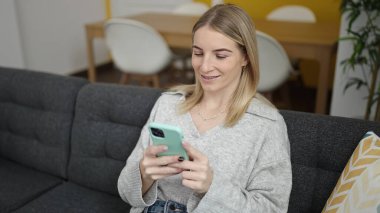 Evdeki kanepede oturan akıllı telefon kullanan genç sarışın kadın.