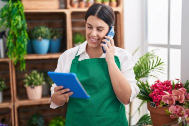 Young beautiful hispanic woman florist talking on smartphone using touchpad at flower shop