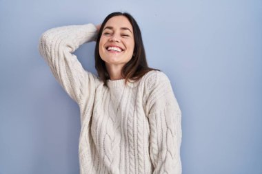 Genç esmer kadın mavi arka planda duruyor. Kendine güvenen, el hareketleriyle saçlara dokunan, çekici ve modaya uygun poz veren. 