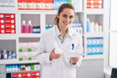 Young woman pharmacist holding pills bottle reading prescription at pharmacy