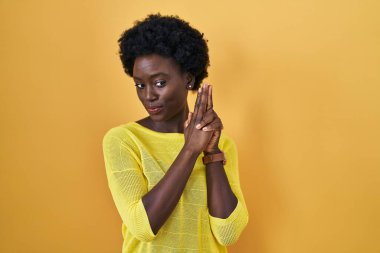 African young woman standing over yellow studio holding symbolic gun with hand gesture, playing killing shooting weapons, angry face 