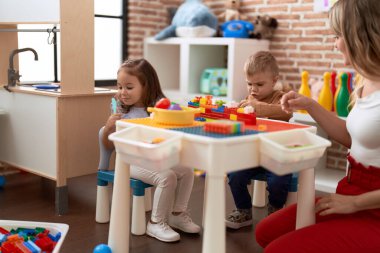 Teacher with boy and girl playing with construction blocks sitting on table at kindergarten
