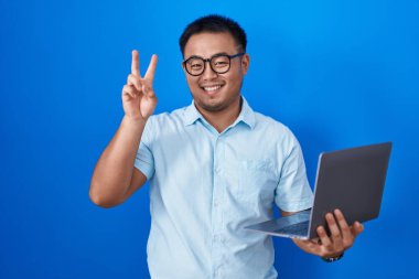 Chinese young man using computer laptop smiling with happy face winking at the camera doing victory sign with fingers. number two. 