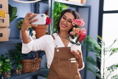 Young beautiful hispanic woman florist make selfie by smartphone holding plant at flower shop