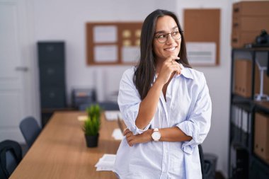 Young hispanic woman at the office with hand on chin thinking about question, pensive expression. smiling and thoughtful face. doubt concept. 