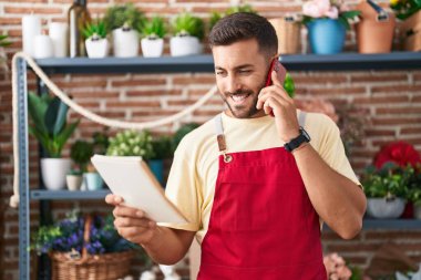 Young hispanic man florist talking on smartphone reading book at florist